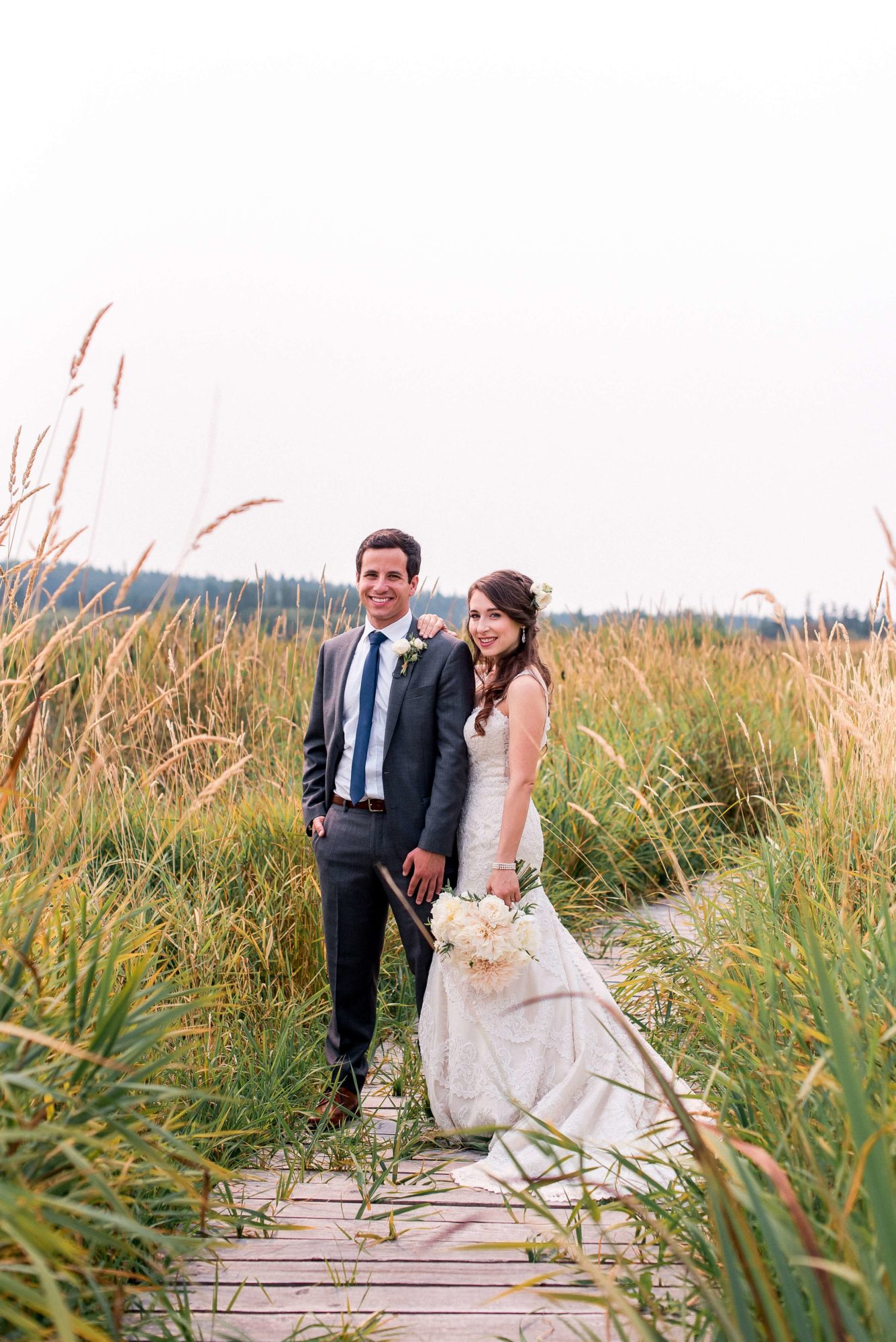 bride and groom stand on overgrown boardwalk in tall field of grain
