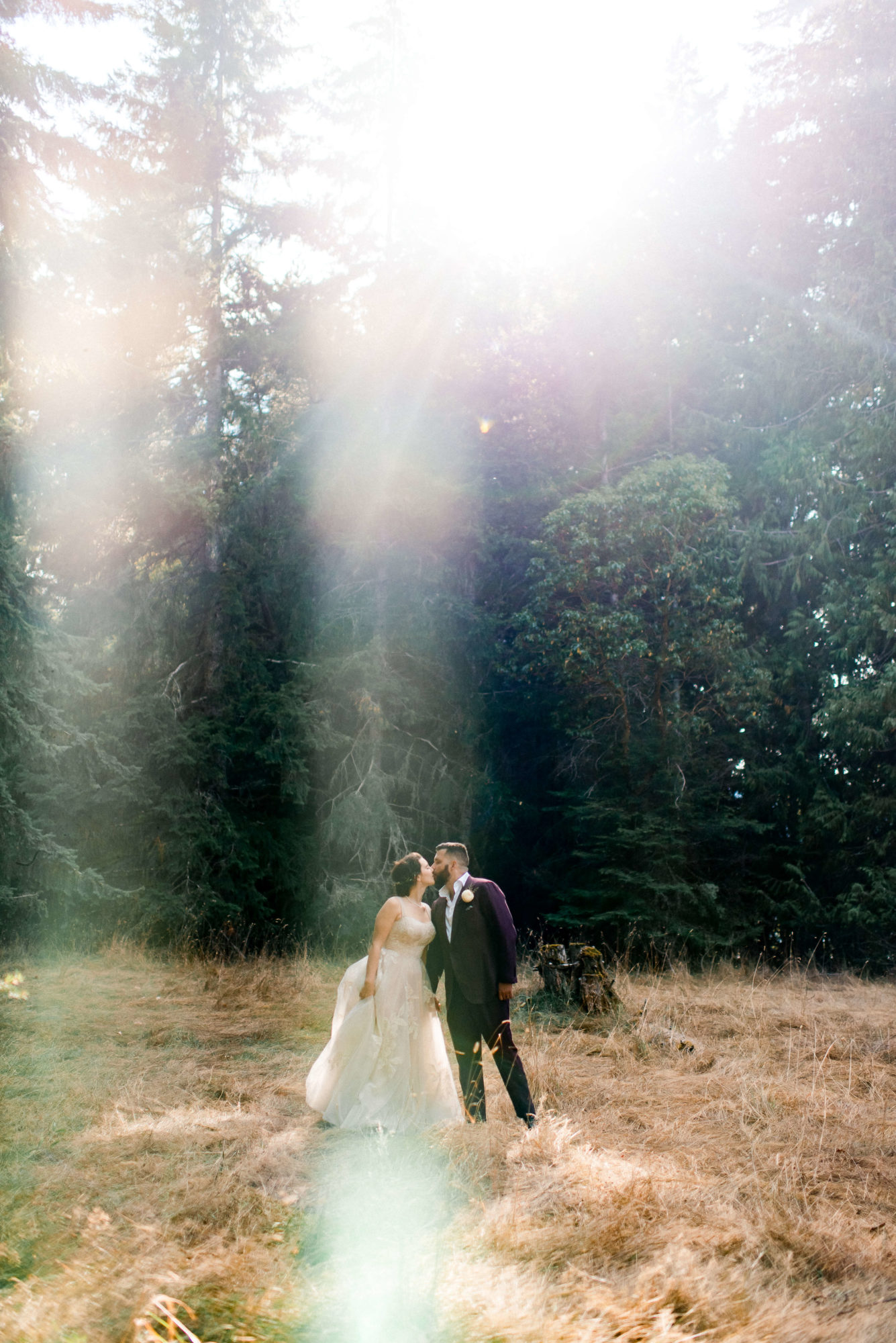bride and groom kiss under sun rays breaking through the forest trees
