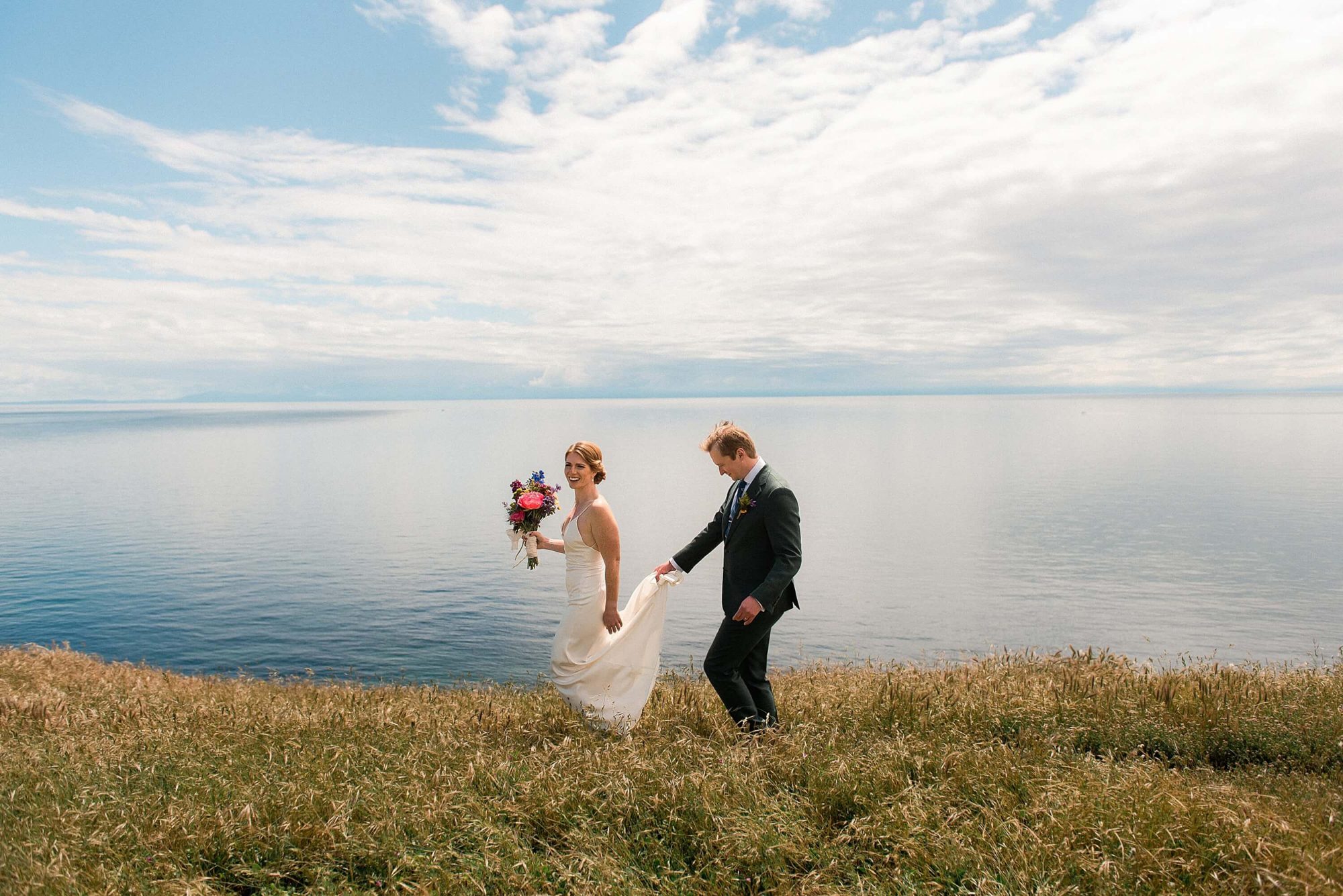 bride and groom walking along a cliffside overlooking the water at their San Juan Island Wedding