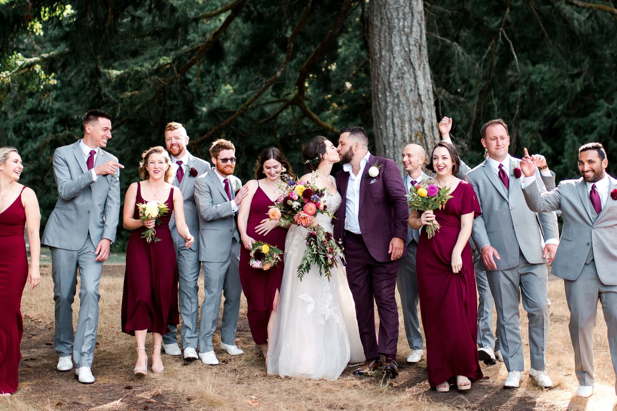 wedding party in grey and maroon in the forest as the bride and groom kiss at Islandwood wedding venue